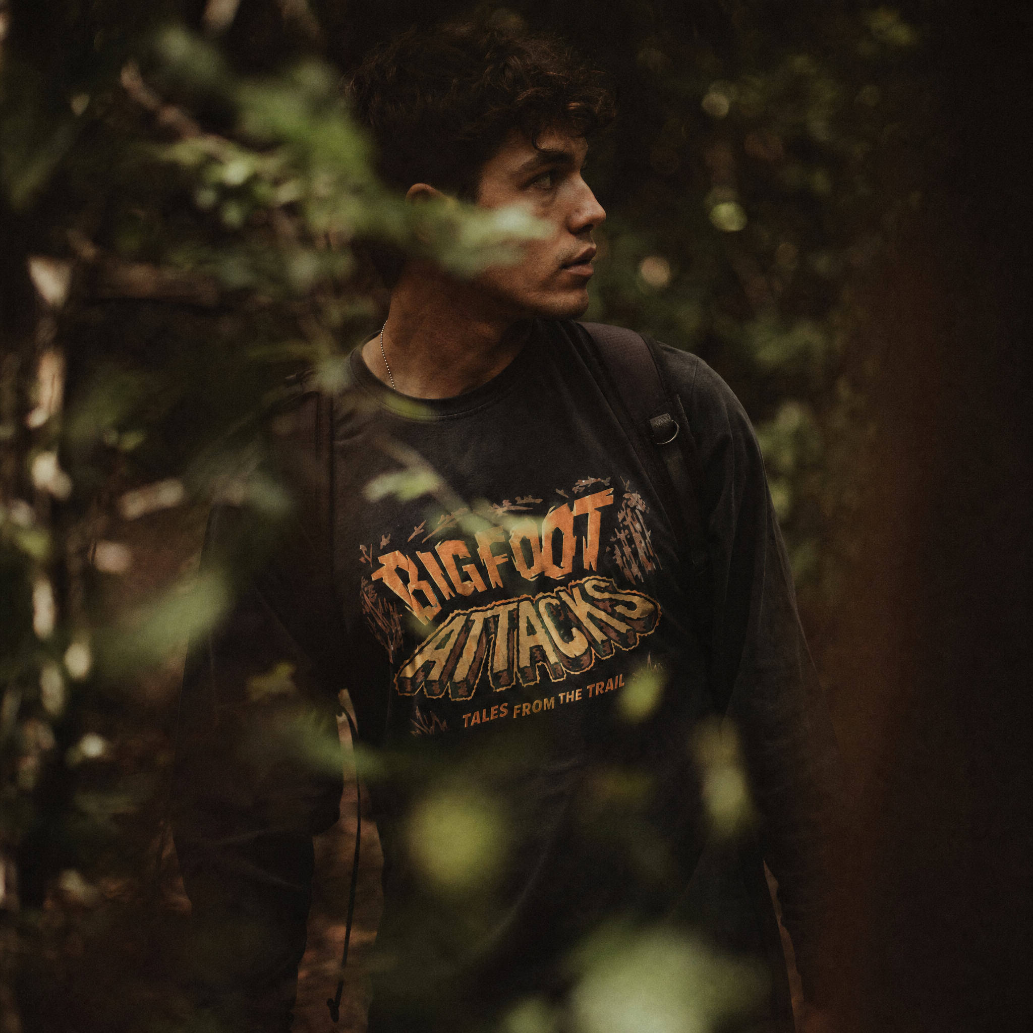 A man wearing a dark graphic t-shirt with 'BIGFOOT ATTACKS' printed on the front, standing in a dark, leafy outdoor forest setting.